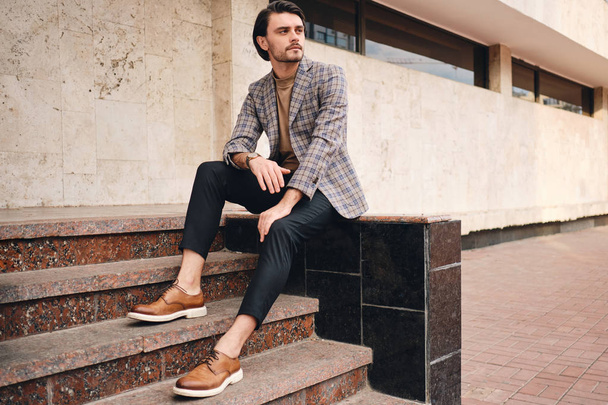 Young handsome stylish man in checkered jacket thoughtfully looking away sitting outdoor - Foto, imagen