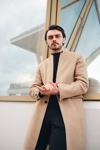 Young serious stylish man in beige coat confidently looking in camera outdoor - Zdjęcie, obraz