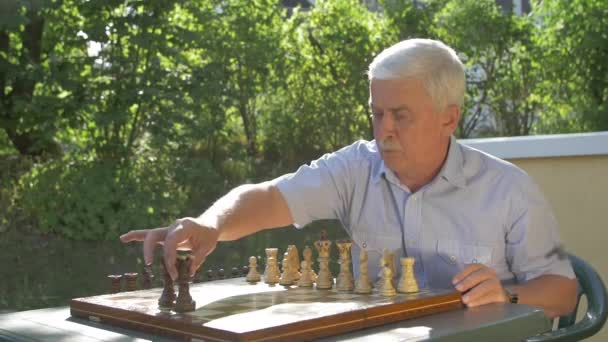 Vecchio uomo dai capelli grigi prepara la partita a scacchi fuori in giardino a tavola e mette le figure nere sulla loro posizione sulla scacchiera
. - Filmati, video
