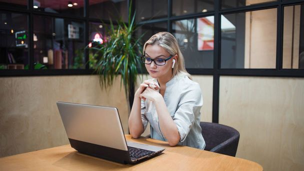 jonge blonde vrouw zit op haar laptop en maakt gebruik van draadloze hoofdtelefoon. - Foto, afbeelding