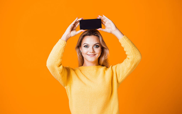 Cute young woman in a yellow sweater holding a smartphone - Zdjęcie, obraz