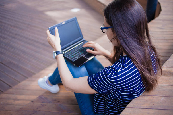 A brunette girl, a student with long hair, with glasses sitting cross-legged on wooden steps and typing on laptop. - Foto, immagini