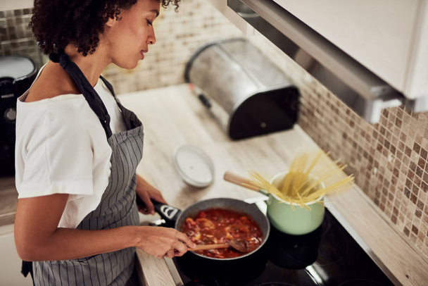 Mixed race woman in apron standing next to stove and stirring toMixed race woman in apron standing next to stove and stirring tomato sauce. On stove are saucepan and pot with spaghetti. - Fotografie, Obrázek