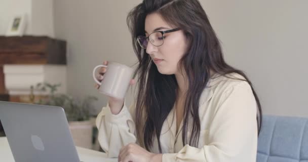 Close-up portrait of a young beautiful Caucasian woman in eyeglasses sitting with laptop and drinking tea or coffee. Intelligent girl with long black hair resting at home. Cinema 4k footage ProRes HQ - Imágenes, Vídeo