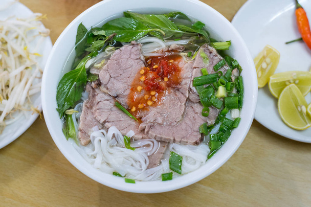 Soup pho in a budget restaurant in Danang city, Vietnam for 35 000 dongs. Noodles, sliced boiled beef, basil leaves, chopped spring onion. Sprouts, chili and pieces of lime on the plate. - Photo, Image