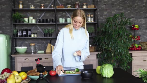 Young beautiful blonde takes on videos as she cooks in the kitchen - Video, Çekim