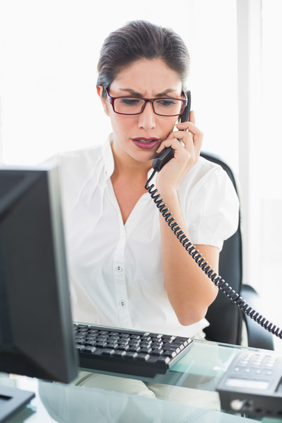 Frowning businesswoman sitting at her desk talking on the phone - Foto, Imagen