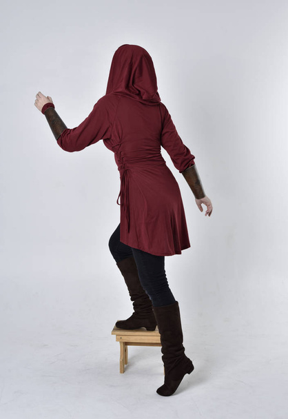 πορτραίτο ενός μελαχρινού κοριτσιού με κόκκινο χιτώνα φαντασίας με κουκούλα. Στέκοντας όρθια στάση σε ένα λευκό φόντο στούντιο. - Φωτογραφία, εικόνα