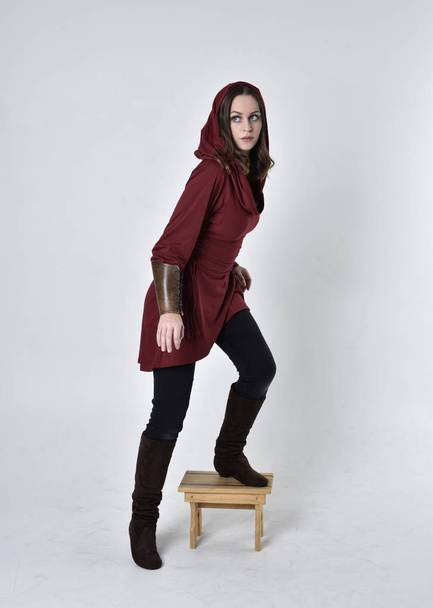 full length portret van een brunette meisje draagt een rode fantasie tuniek met kap. Stepping up staande houding op een witte studio achtergrond. - Foto, afbeelding