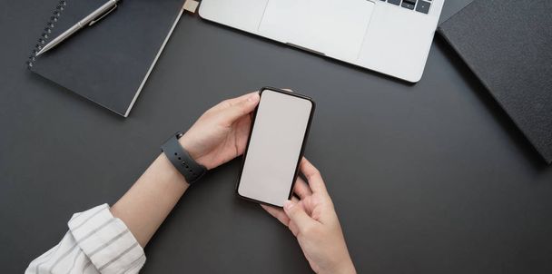 Fotografía aérea de una joven empresaria sosteniendo un teléfono inteligente de pantalla en blanco en un espacio de trabajo de lujo oscuro con suministros de oficina
  - Foto, imagen