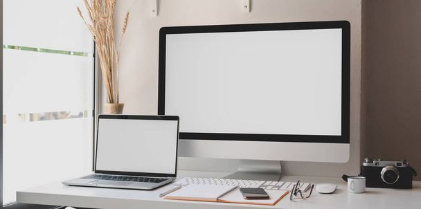Blank screen desktop computer and laptop in modern workspace with office supplies  - 写真・画像
