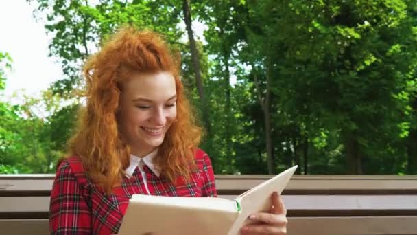 Jovial red haired girl laughing at funny book in park - Video