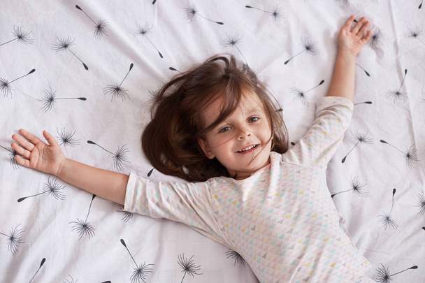 Cute little girl playing at home while lying on blanket with dandelion on bed with sleepy and happy facial expression, spreading her arms. Sweet elementary age child having fun in cosy bedroom. - Photo, image