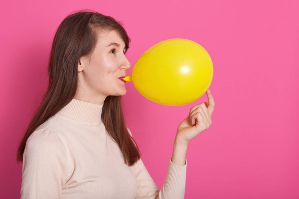 Side view of funny young woman with long dark hair inflating yellow balloon isolated over pink background, adorable female wearing rosy shirt, copy space for your advertisment or promotion text. - Photo, image