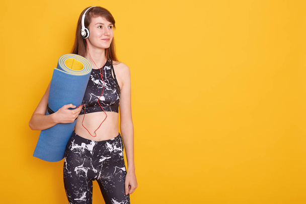 Young athletic woman wearing headphones and black sportswear holding blue yoga mat in hands, looking aside, ready for doing sport, isolated over yellow wall, copy space for promotion or advertismant. - Φωτογραφία, εικόνα