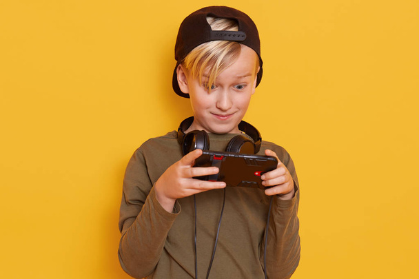 Portrait of emotional and active little boy with blond hair and green shirt, carrying his finger on screen of smartphone while playing his favorite online game, poses over yellow background in studio. - Фото, зображення