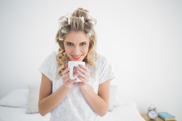Smiling cute blonde wearing hair curlers smelling coffee - Фото, изображение