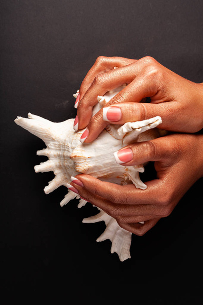 Esmalte de gel de manicura francés. Concepto de uñas fuerte y saludable. Hermosas manos femeninas sobre un fondo negro con un cockleshell
. - Foto, imagen