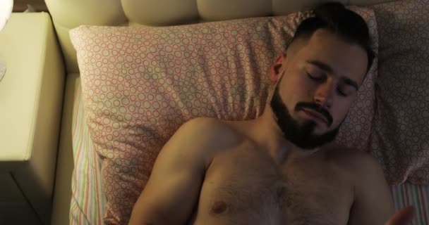 A young man with a beard is waking up early in the morning and turning on a lamp or night light. - Video