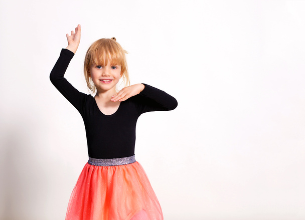 Klein blond ballerina meisje poseert in een dansclub met houten vloer en witte muur. Jonge balletdanser in roze tutu, die plezier heeft en lacht. Achtergrond, kopieer ruimte. - Foto, afbeelding