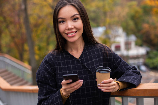 Joven hermosa mujer en abrigo con café para llevar y teléfono celular mirando felizmente en la cámara en el parque
 - Foto, imagen