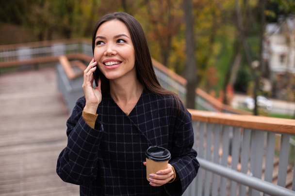 Joven mujer alegre casual en abrigo con café para ir felizmente hablando por teléfono celular en el parque
 - Foto, Imagen