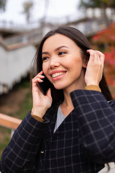 Portrait of young cheerful woman in coat happily talking on cellphone in city park - Photo, Image