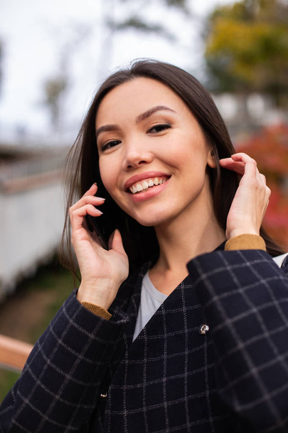 Portret van jonge mooie vrouw in jas blij kijkend in camera pratend op mobiele telefoon in stadspark - Foto, afbeelding