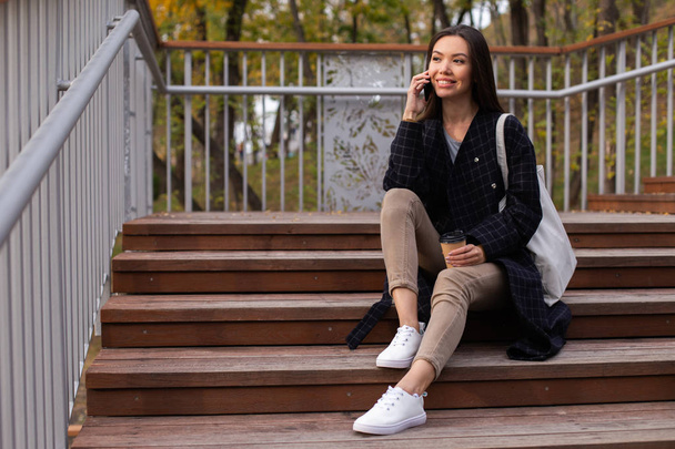 Fiatal gyönyörű alkalmi nő kávé menni boldogan beszél mobilon a lépcsőn a parkban - Fotó, kép