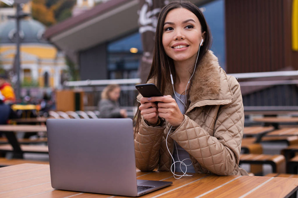 junge schöne, lässige Frau in Daunenjacke mit Laptop benutzt fröhlich Handy in Café auf der Straße - Foto, Bild