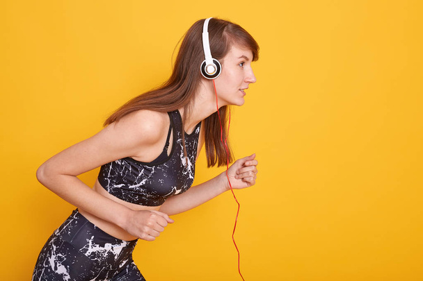 Portrait of fit and sporty young brunette woman preparing for running, listening music with white headphones, female wraring blacksport top and leggins, posing isolated over yellow studio concept. - 写真・画像