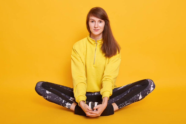 Hoprizontal shot of attractive young woman meditating while sitting on floorin studio, posing isolated over yellow background, female wearing hoody and leggins. Sport, fitness and lifestyle concept. - Zdjęcie, obraz