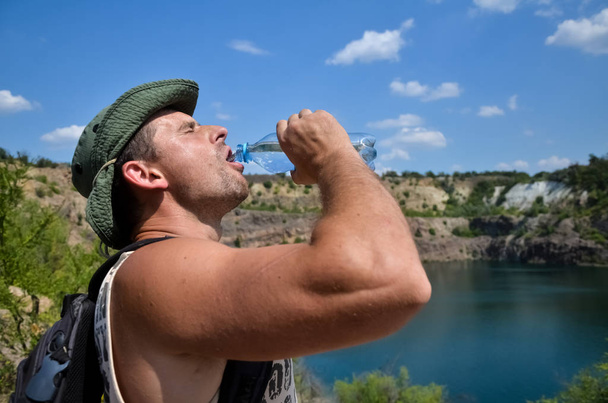 Un joven con gotas de sudor en la cara, en un sombrero y con una mochila bebe agua fresca y limpia de una botella de plástico en un caluroso día de verano contra un cielo azul y un lago de agua dulce
. - Foto, imagen