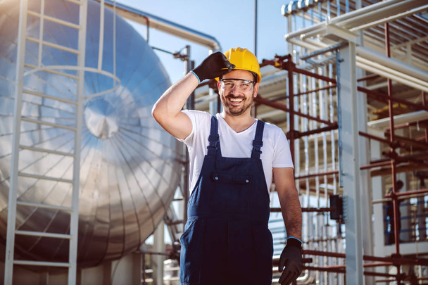 Happy caucasian worker in overall and with helmet on head posing in front of oil storage tank. - Photo, Image