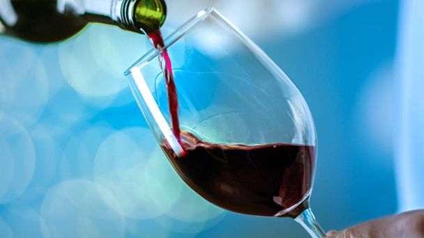 Человек, вливающий красное вино в бокал вина. Фон синий с размытыми огнями
. - Фото, изображение