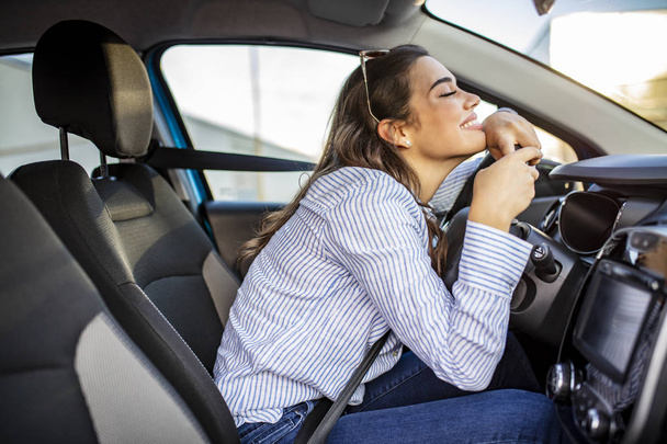 Young Woman Embracing Her New Car. Excited young woman and her new car indoors. Young and cheerful woman enjoying new car hugging steering wheel sitting inside - Foto, Bild