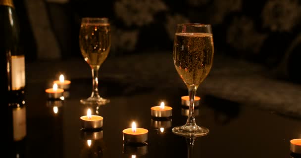 Full glasses of Champagne on black glossy table with red burning candles and champagne bottle, women sitting on sofa in background. Cosy romantic Christmas vacation atmosphere - Materiał filmowy, wideo