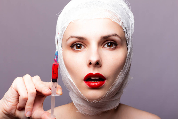 beautiful young girl, lips painted up with red lipstick, head bandaged with a bandage, in the hands of a syringe with a red liquid - Foto, Imagen