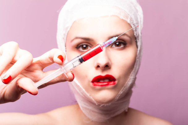 beautiful young girl, lips painted up with red lipstick, head bandaged with a bandage, in the hands of a syringe with a red liquid - Foto, imagen