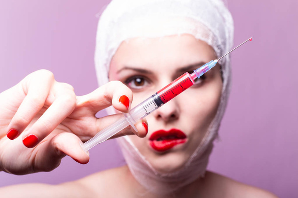 beautiful young girl, lips painted up with red lipstick, head bandaged with a bandage, in the hands of a syringe with a red liquid - Foto, Bild