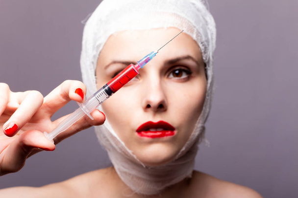 beautiful young girl, lips painted up with red lipstick, head bandaged with a bandage, in the hands of a syringe with a red liquid - Foto, Bild