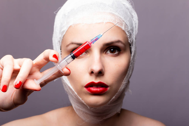 beautiful young girl, lips painted up with red lipstick, head bandaged with a bandage, in the hands of a syringe with a red liquid - Photo, Image