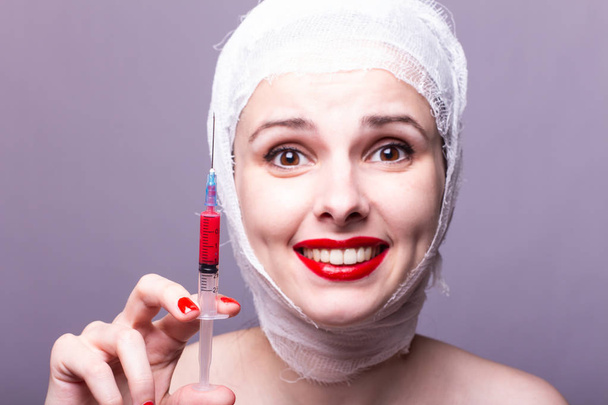beautiful young girl, lips painted up with red lipstick, head bandaged with a bandage, in the hands of a syringe with a red liquid - Foto, afbeelding