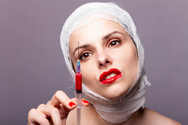 beautiful young girl, lips painted up with red lipstick, head bandaged with a bandage, in the hands of a syringe with a red liquid - Photo, Image