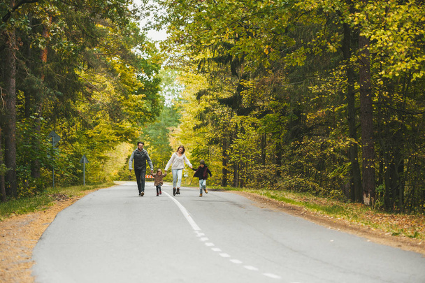 Η μαμά, ο μπαμπάς και τα μικρά παιδιά, περπατούν σε επαρχιακό δρόμο. Μιλάνε και απολαμβάνουν μια όμορφη φθινοπωρινή μέρα.. - Φωτογραφία, εικόνα
