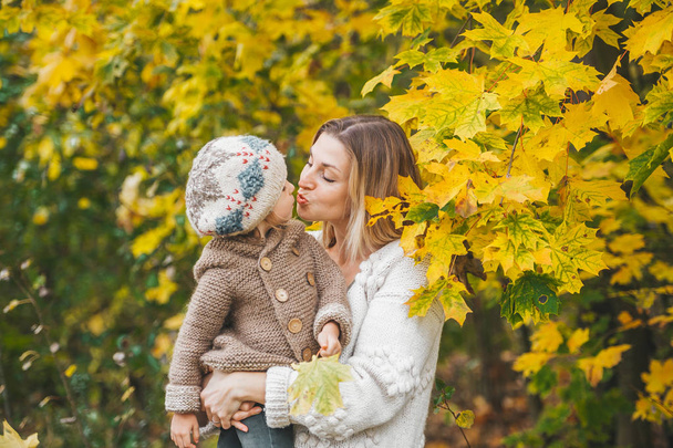Madre besándose y abrazando a su hermosa hija pequeña mientras está de pie cerca del árbol de arce en el parque de otoño
. - Foto, Imagen