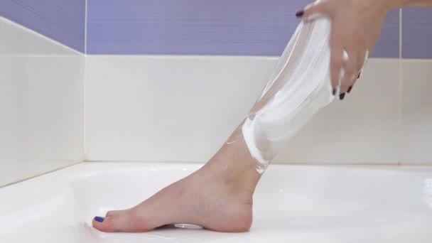 Μια νεαρή γυναίκα ξυρίζει τρίχες στα πόδια της. Το κορίτσι έβαλε αφρό ξυρίσματος στο πόδι της και άρχισε να ξυρίζεται.. - Πλάνα, βίντεο