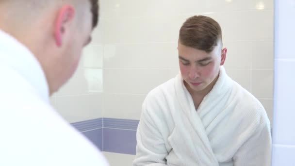 A young man puts cosmetic patches on his face. Spa treatments. Brown hydrogel patches for skin rejuvenation. He is standing in a white bathrobe in front of the bathroom mirror. View through the mirror - Video