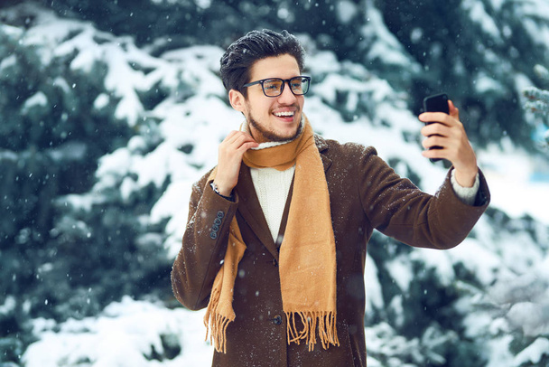 Young man in a coat on the street with a phone. Smiling young man having fun outdoors. Businessman uses a phone.  Man enjoy a winter. Dressed in a coat, sweater and scarf. Winter concept. Snowfall. - Foto, Bild