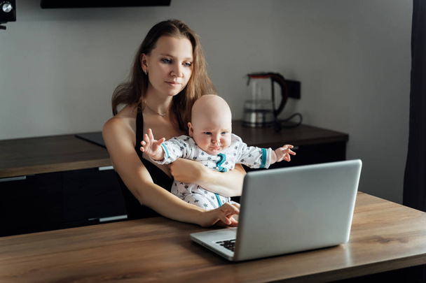 Young mother sits at a table with a baby in her arms and works o - Фото, изображение
